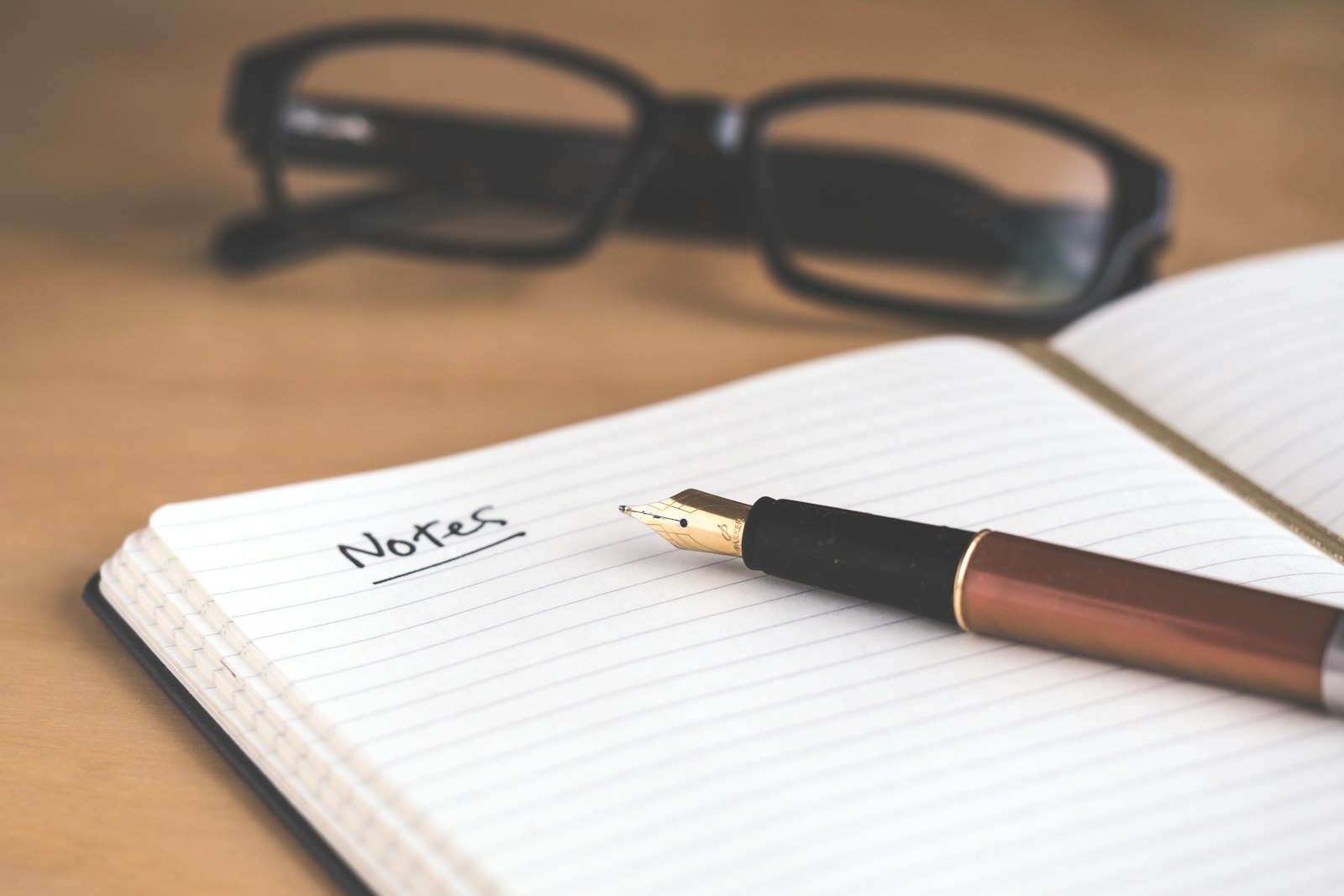 A fountain pen resting on an open notebook with handwritten ‘Notes’ underlined, reading glasses in soft focus behind.